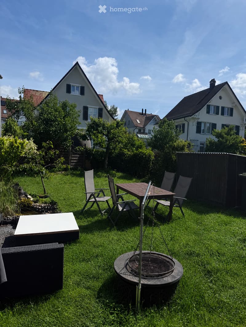 Garden / Outdoor Area. Well-maintained garden with a wooden table, four chairs, a fire pit, and a black outdoor sofa. The area is surrounded by greenery and trees, with residential houses visible in the background.
