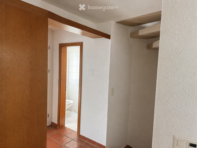 Corridor / Hallway. A narrow hallway with wooden doors, white walls, and built-in wooden shelves. An open doorway leads to a small bathroom with white tiles and a toilet.