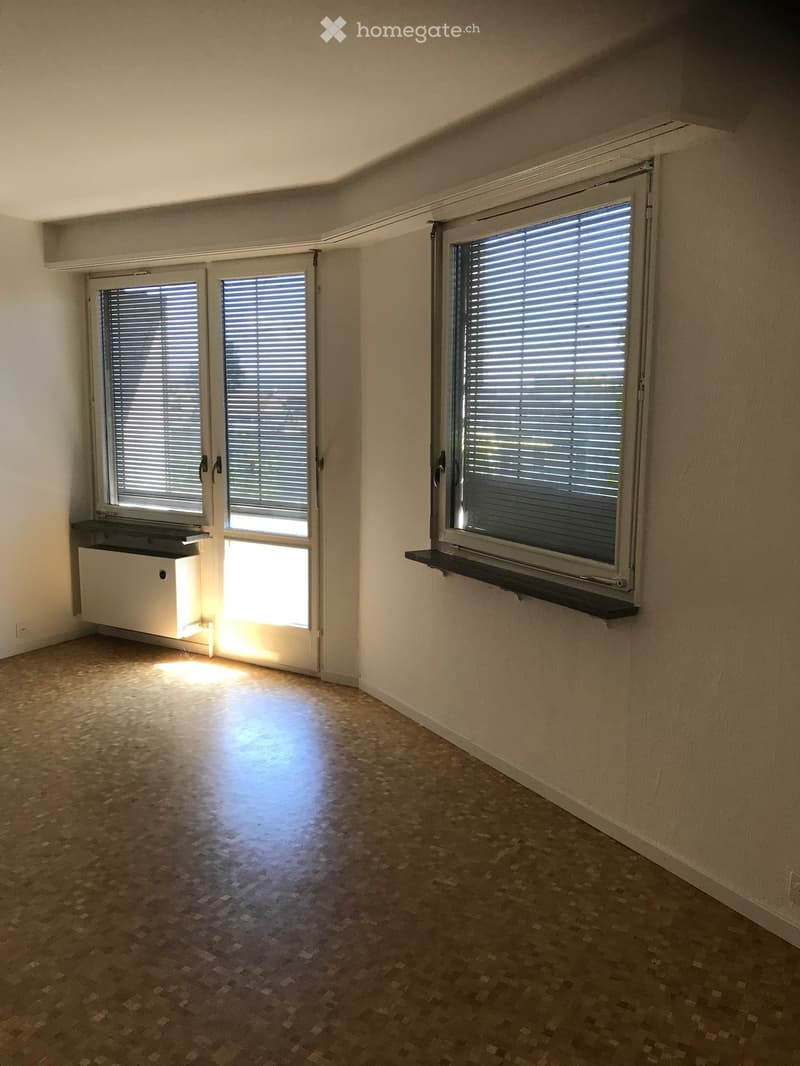 Empty room with wooden flooring, large windows with blinds, and a small shelf on the right side.