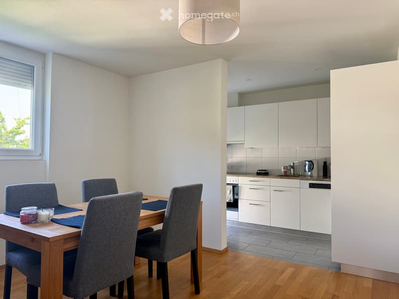 Open-plan dining and kitchen area with a wooden dining table, four grey chairs, white cabinets, and a modern kitchen setup including an oven and dishwasher.