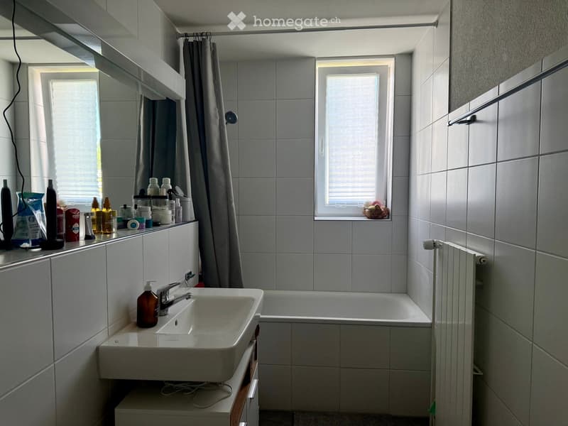 Modern bathroom with a white tiled wall, a sink with a faucet, a bathtub, a mirror, and a cabinet with toiletries. There is a window with blinds and a radiator on the wall.