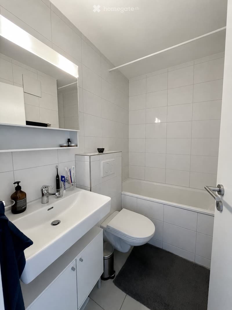 Modern bathroom with white tiles, a sink with a faucet, a toilet, a bathtub, a mirror, and a small cabinet. The room includes a toothbrush holder, soap dispenser, and a towel on the floor.