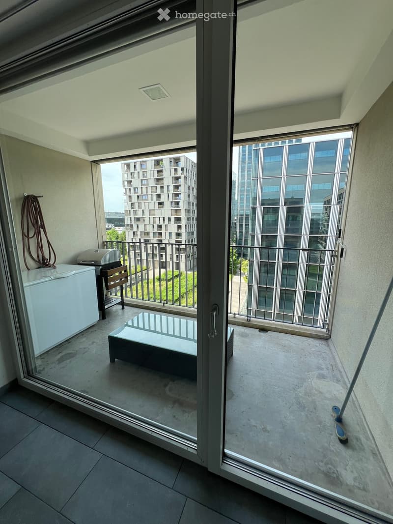 Balcony with a glass door, a small table, a chair, a refrigerator, and a grill. The view shows modern high-rise buildings.