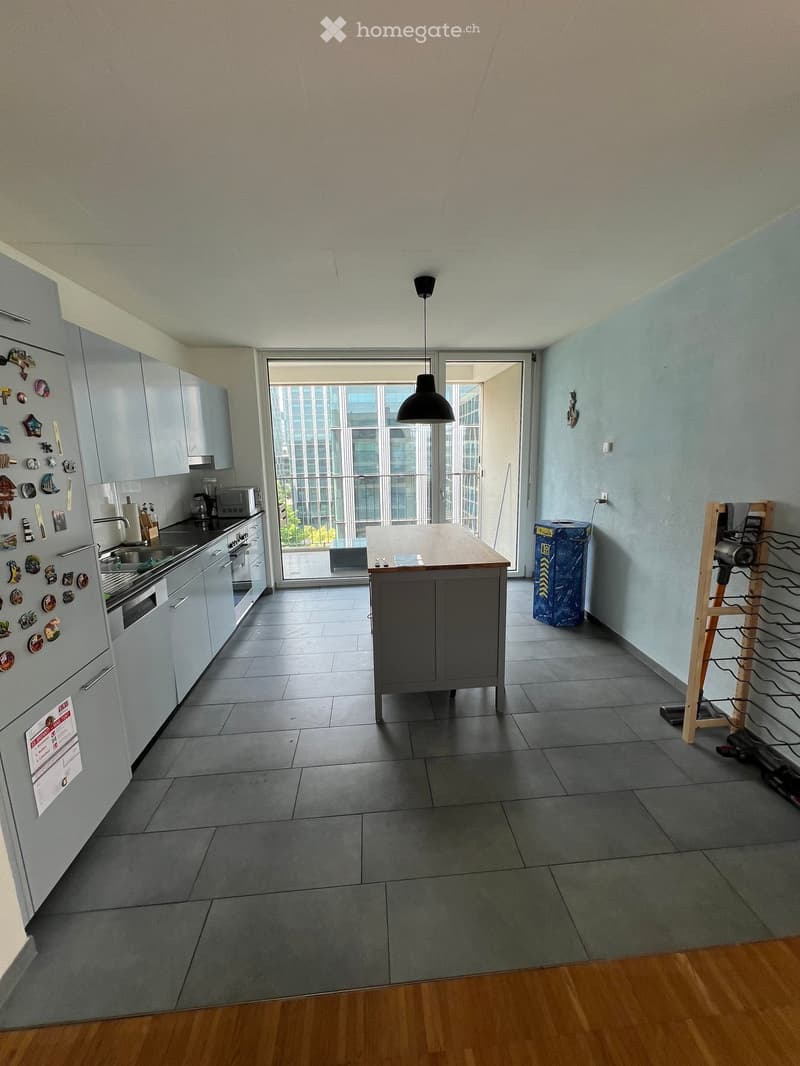 Modern kitchen with grey cabinets, stainless steel appliances, a central island, and a view of the city through large windows.