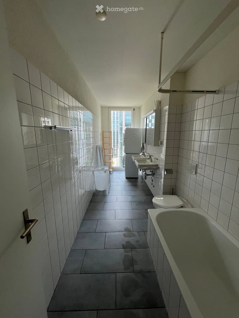 Modern bathroom with tiled walls and floor, featuring a bathtub, toilet, sink, and mirror. The space is well-lit with natural light from a large window at the end of the room.