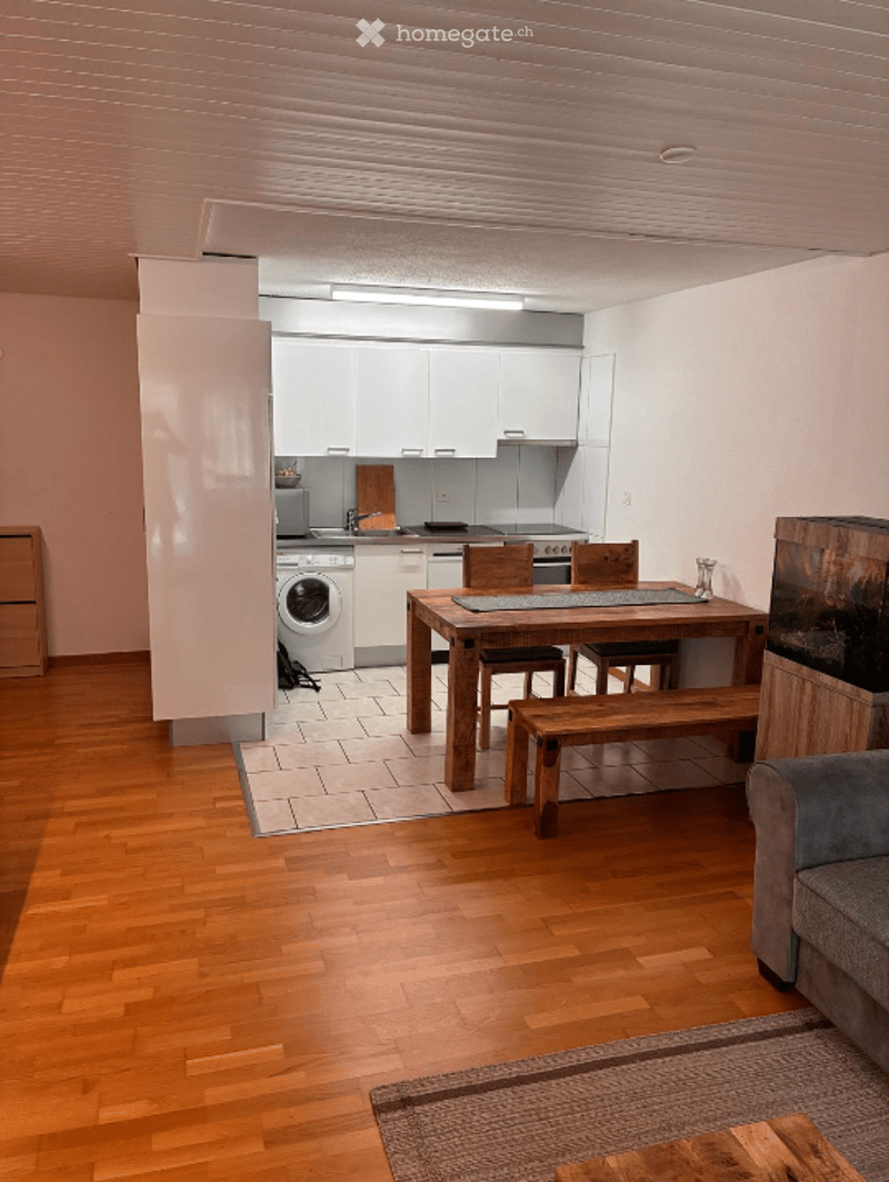 Open-plan living and dining area with a sofa, dining table, bench, chairs, and a small kitchen with a washing machine, fridge, and oven.
