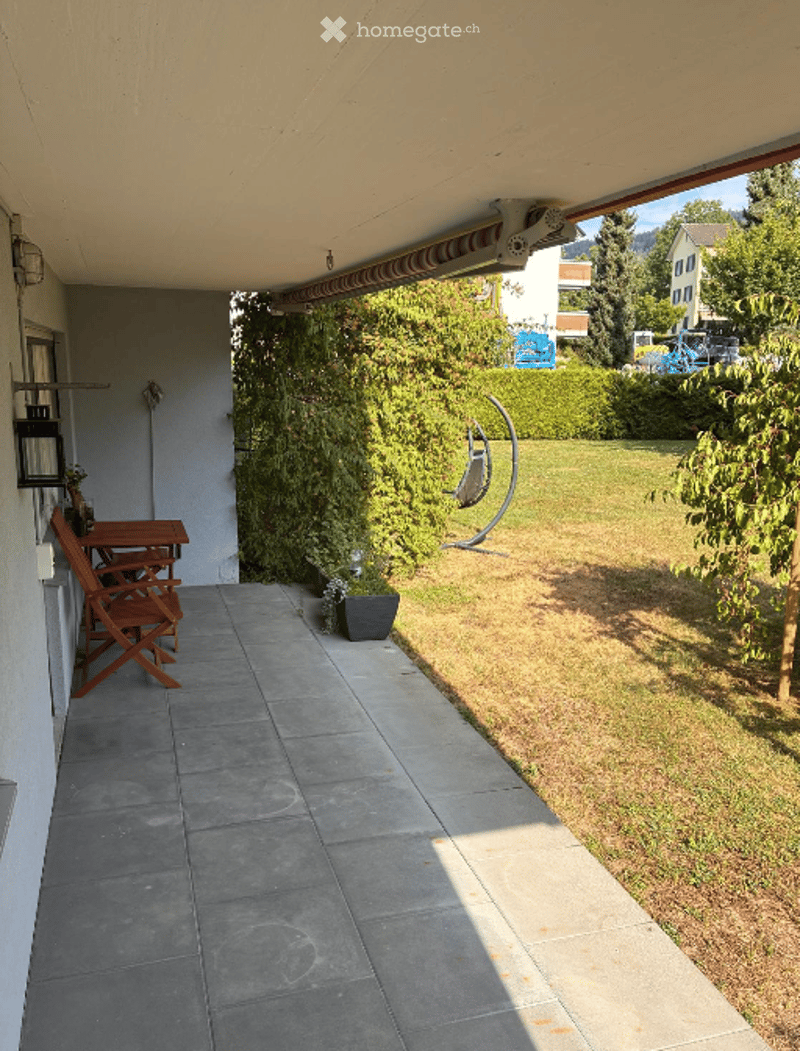 Balcony with tiled floor, wooden table and chairs, potted plant, and a view of a green lawn and garden area.