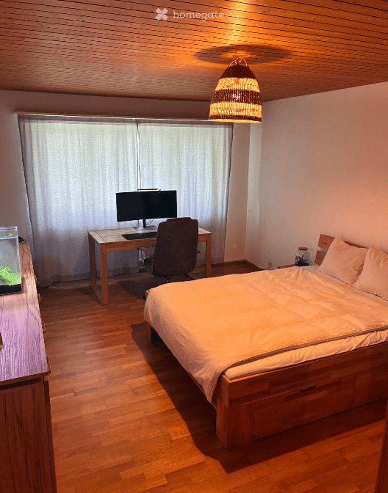 Cozy bedroom with a wooden bed, desk with a computer, chair, and a small fish tank on a side table. The room features wooden flooring and a hanging woven light fixture.