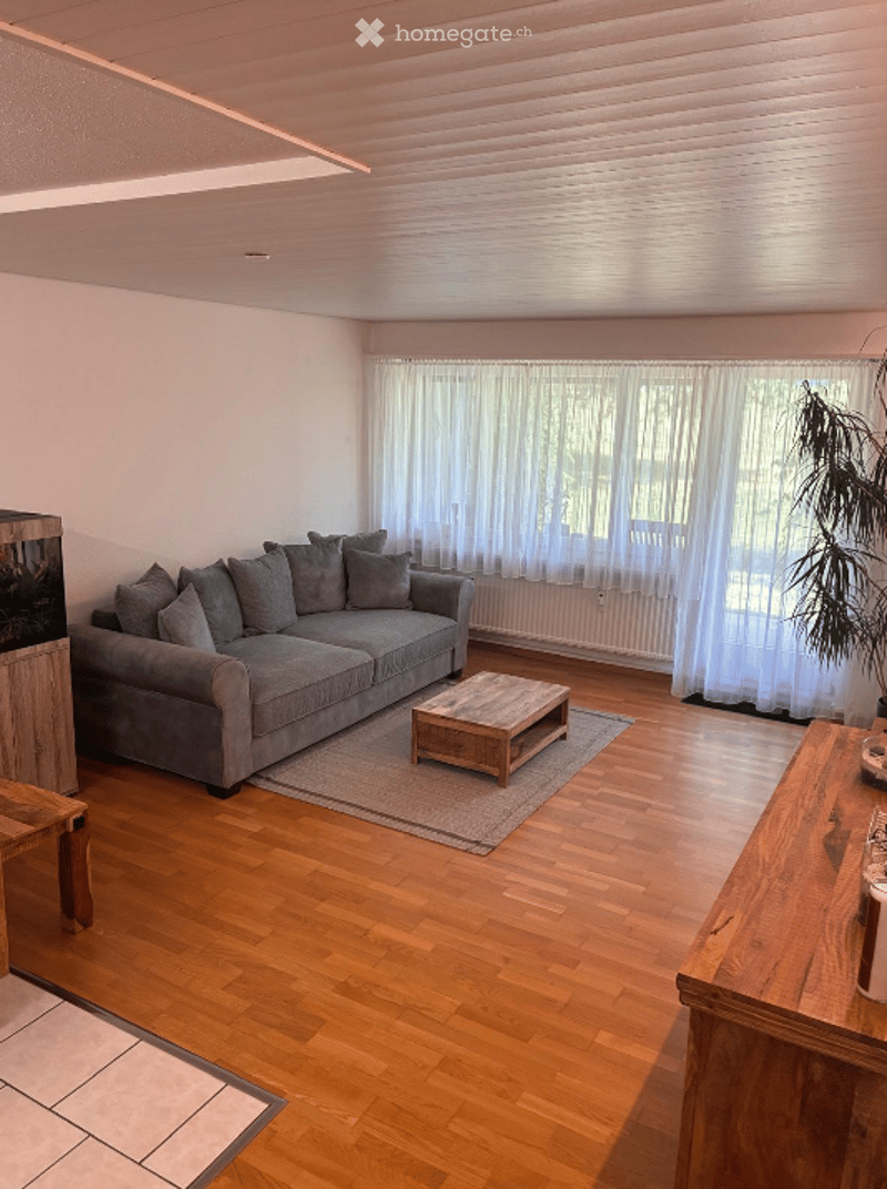 Bright living room with wooden flooring, grey sectional sofa, coffee table, and large windows with white curtains.