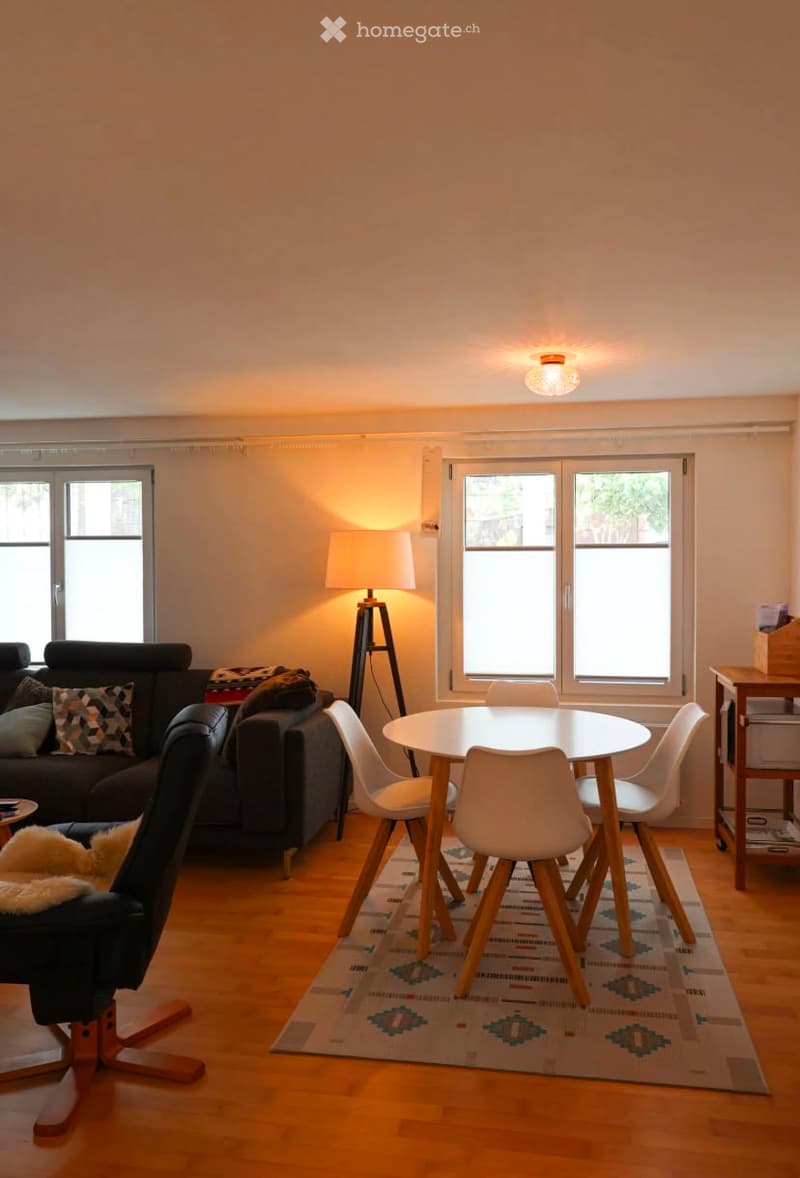 Bright living room with wooden flooring, large windows, a sofa, a round dining table with chairs, a floor lamp, and an ottoman.