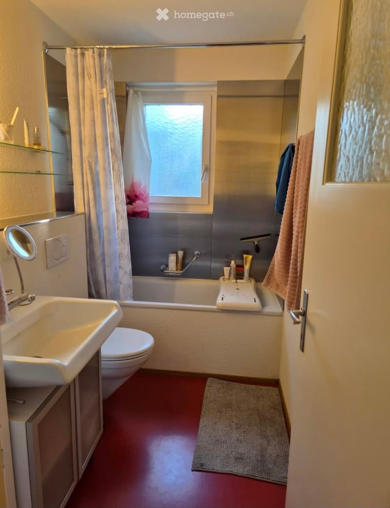 Bathroom with red floor, white sink, toilet, bathtub, mirror, and towel. Features a window above the bathtub with a shower curtain.