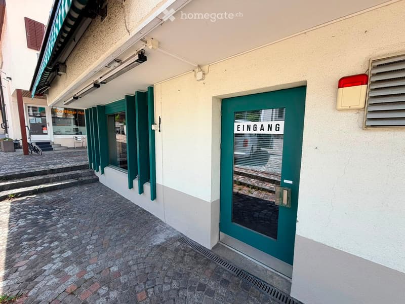 Exterior view of a building entrance with a green door labeled 'EINGANG' and a small staircase leading to a neighboring storefront.
