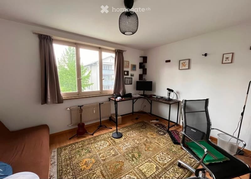 A cozy, well-lit room with a large window, a patterned rug, a brown sofa, a desk with a computer, a guitar on a stand, and a modern hanging light fixture.