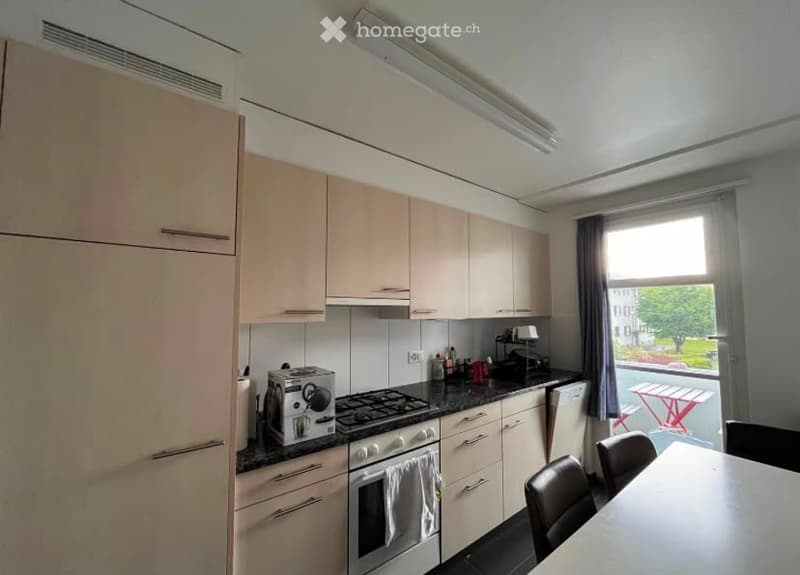 Modern kitchen with light-colored cabinets, stainless steel appliances, and a black countertop. Includes a refrigerator, oven, stove, dishwasher, toaster, coffee maker, and a small dining area with a table and chairs.