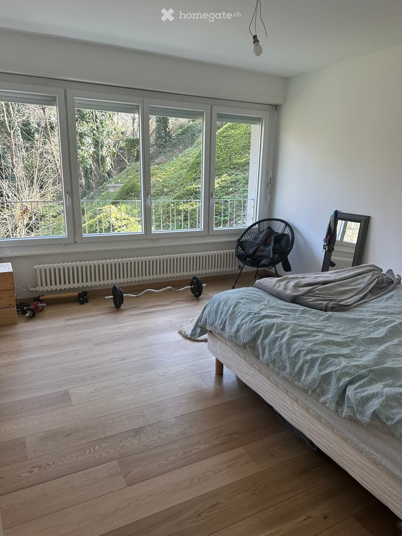 Bright bedroom with wooden flooring, large windows offering a view of greenery, a bed with a blue duvet, a black chair with a jacket, a mirror, a radiator, and a weight bench with weights.