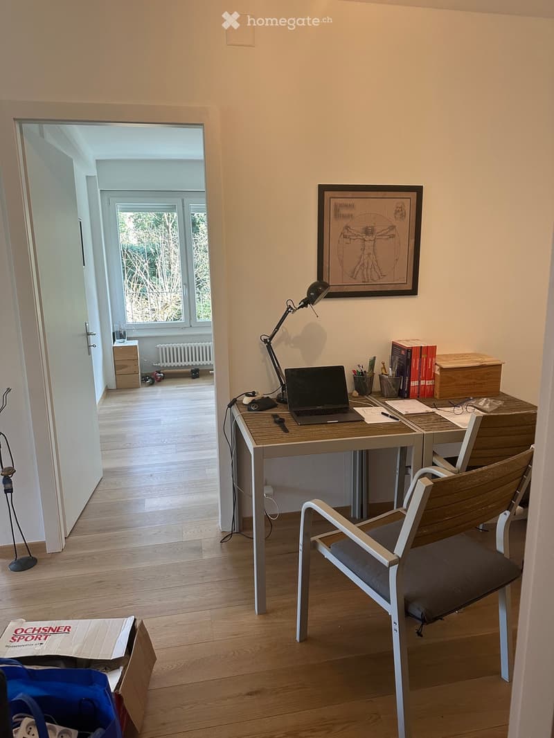 A modern, well-lit room with a wooden floor, a desk with a laptop, a chair, a lamp, and a framed picture on the wall. The room has an open doorway leading to another space with large windows and a radiator.