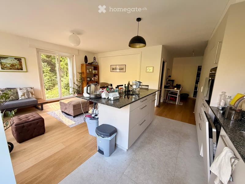 Open-plan living and kitchen area with a grey sofa, ottoman, and a kitchen island with various appliances and items on the counter. The space features wooden flooring, large windows with a view of greenery, and modern kitchen cabinets.