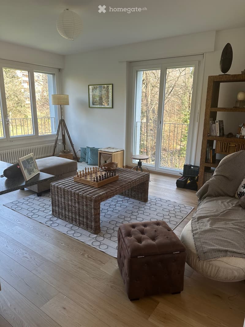 Bright living room with wooden flooring, large windows, and a balcony. Features a wicker coffee table with a chessboard, a brown ottoman, a grey sofa, a bookshelf, and a floor lamp.