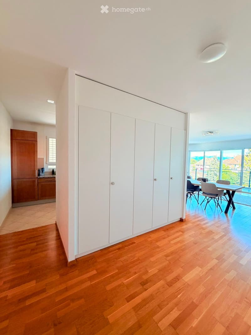 Bright hallway with wooden flooring, white built-in storage units, and a view of a dining area with a table and chairs through an open doorway.