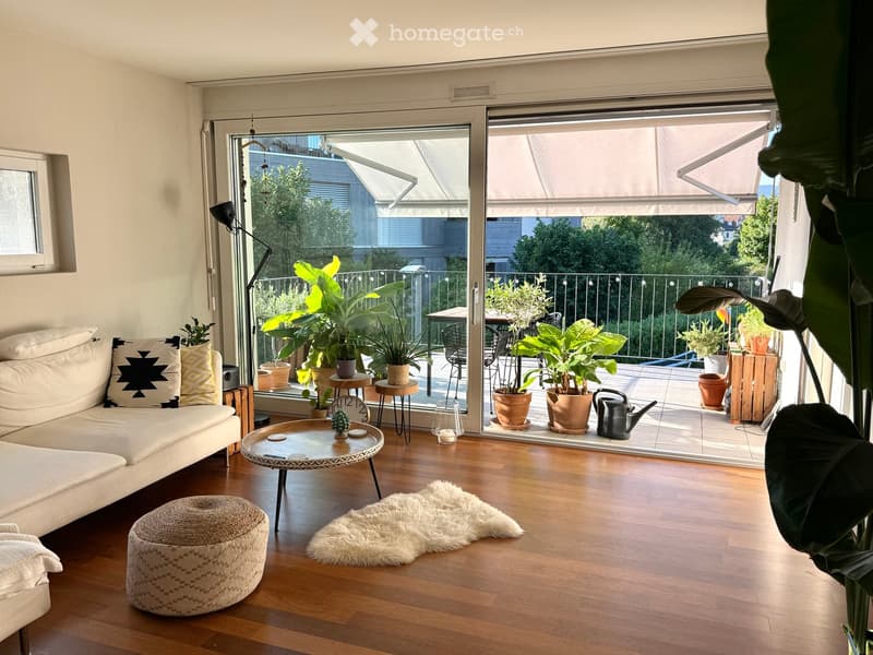 Bright living room with wooden flooring, white sofa, coffee table, and a large sliding glass door leading to a balcony with potted plants and outdoor furniture.