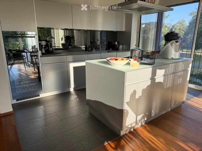 Modern kitchen with white cabinets, a central island, and large windows offering a scenic view. The island features a bowl of fruit and a coffee maker, while the kitchen includes a sink, fridge, and a small dining area with a chair and table.