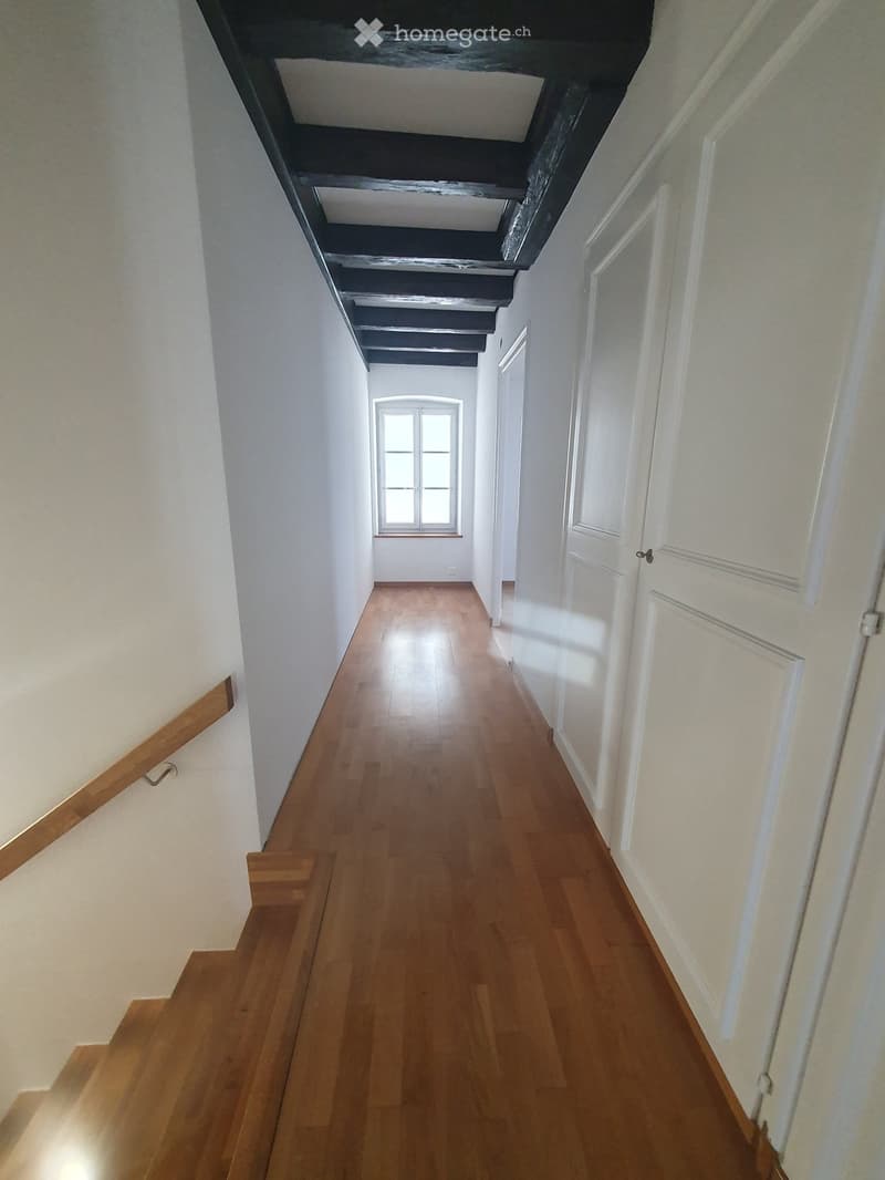 A long, narrow hallway with wooden flooring, white walls, and several closed white doors on the right side. A window at the far end allows natural light to enter.