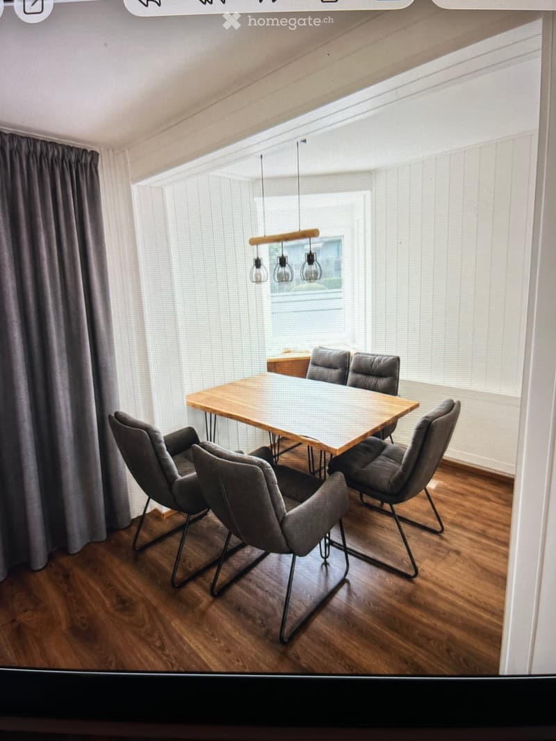 Modern dining area with a wooden dining table, six grey chairs, pendant lights, and large windows with blinds.