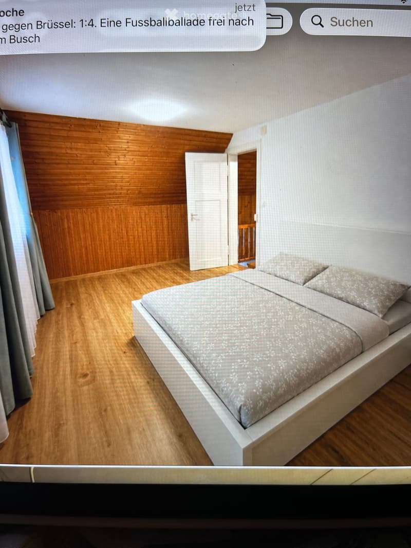 Modern bedroom with a neatly made bed, wooden flooring, and a wooden wall panel. A door leads to another room.