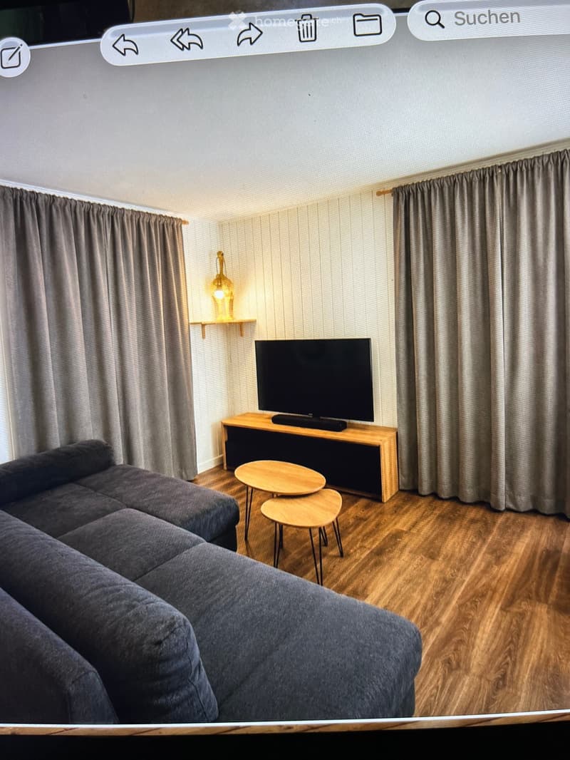 A modern living room with a grey sectional sofa, a wooden coffee table, a flat-screen TV on a wooden stand, and a small round wooden table with a lamp on a shelf.