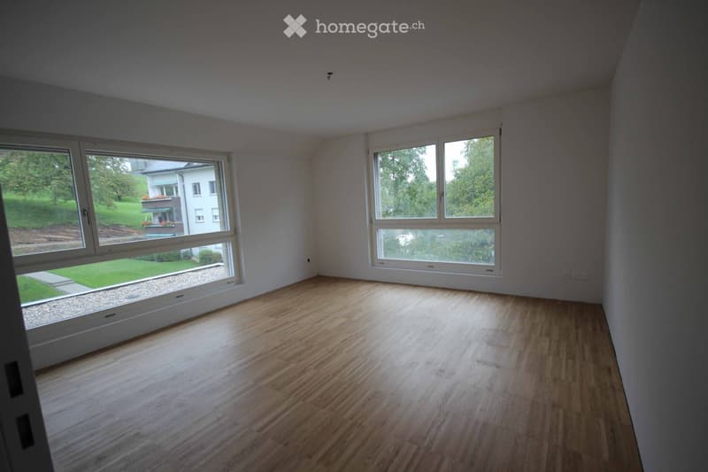 Empty room with wooden flooring and large windows offering a view of a green outdoor area and neighboring house.