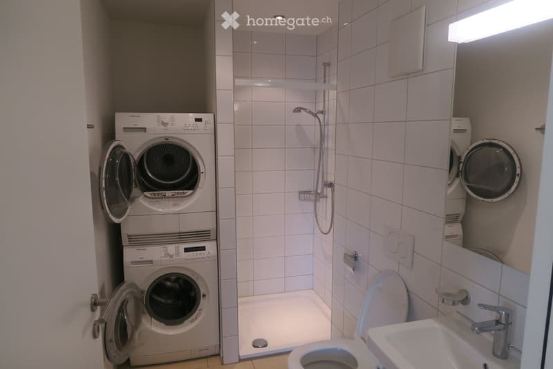 Modern bathroom with white tiled walls, a toilet, sink, shower, and a stacked washing machine and dryer unit.