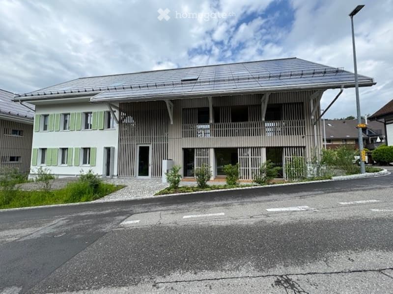 Modern two-story building with a flat roof, green shutters, and a covered balcony area. The entrance features a small garden with low plants and shrubs.
