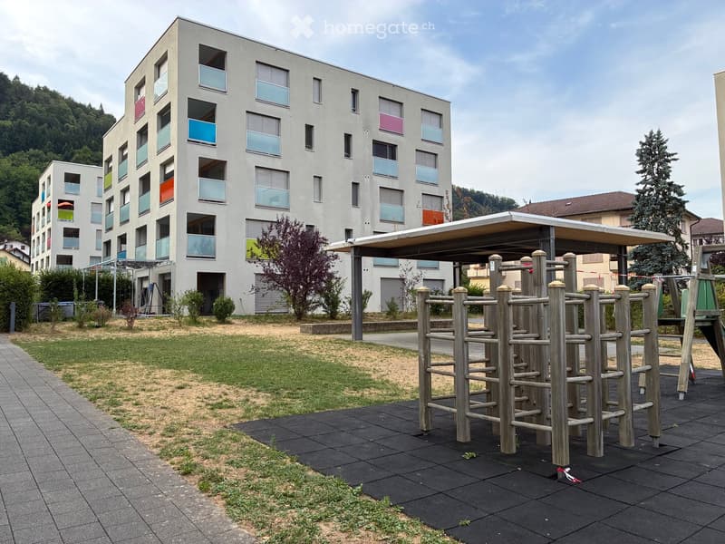 Modern residential building with colorful window frames, surrounded by a grassy area and playground equipment including a wooden climbing structure.