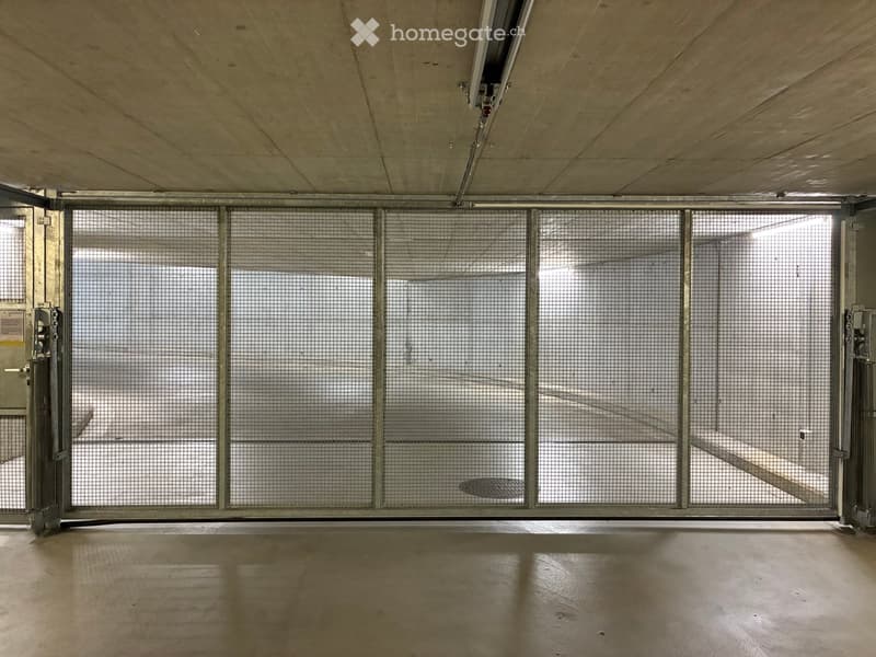Empty underground parking area with concrete floors, metal security gates, and a raised ceiling with lighting fixtures.