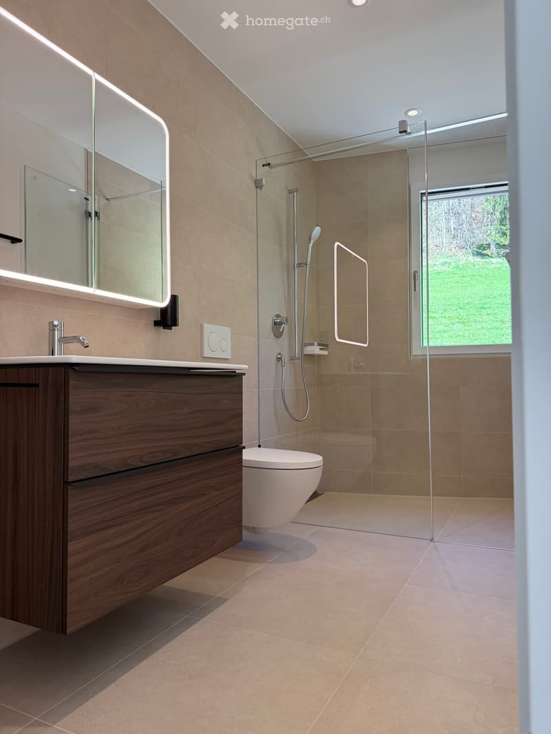 Modern bathroom with beige tiles, wooden vanity, sink, mirror, toilet, and glass-enclosed shower with a modern showerhead.