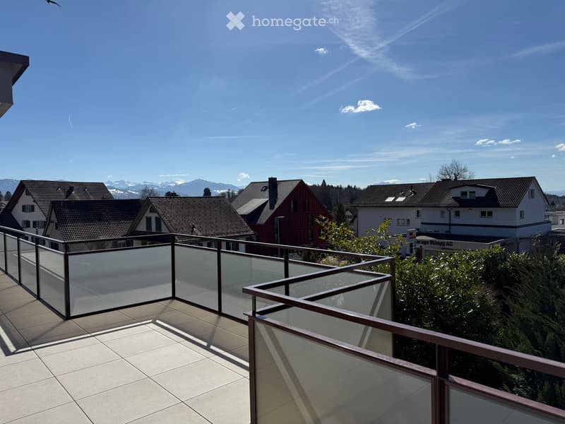 Balcony with glass railing, tiled floor, overlooking a residential area with houses and mountains in the background.