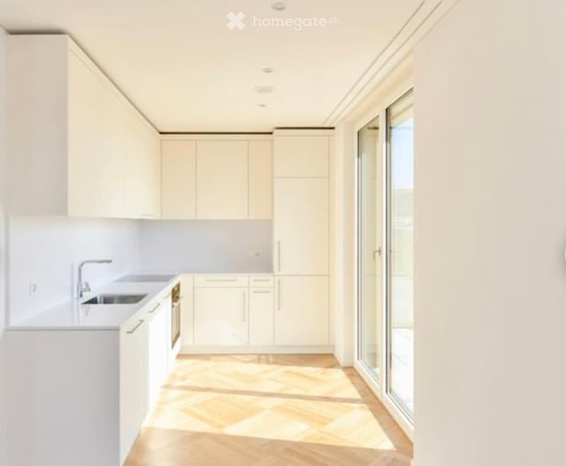 Modern, minimalist kitchen with white cabinets, a central island, and a large window letting in natural light. The floor is light wood, and the space is uncluttered and bright.