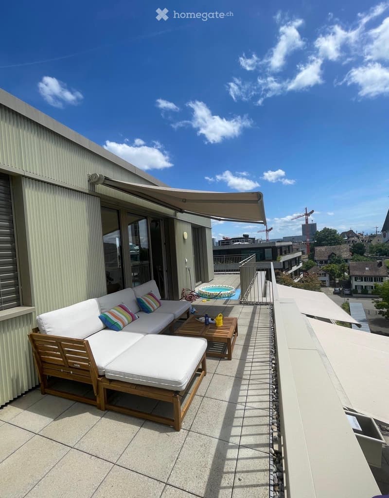 Balcony with a white sofa, wooden coffee table, ottoman, and small round table with a yellow bucket and other items. There is also a small inflatable pool visible in the background.