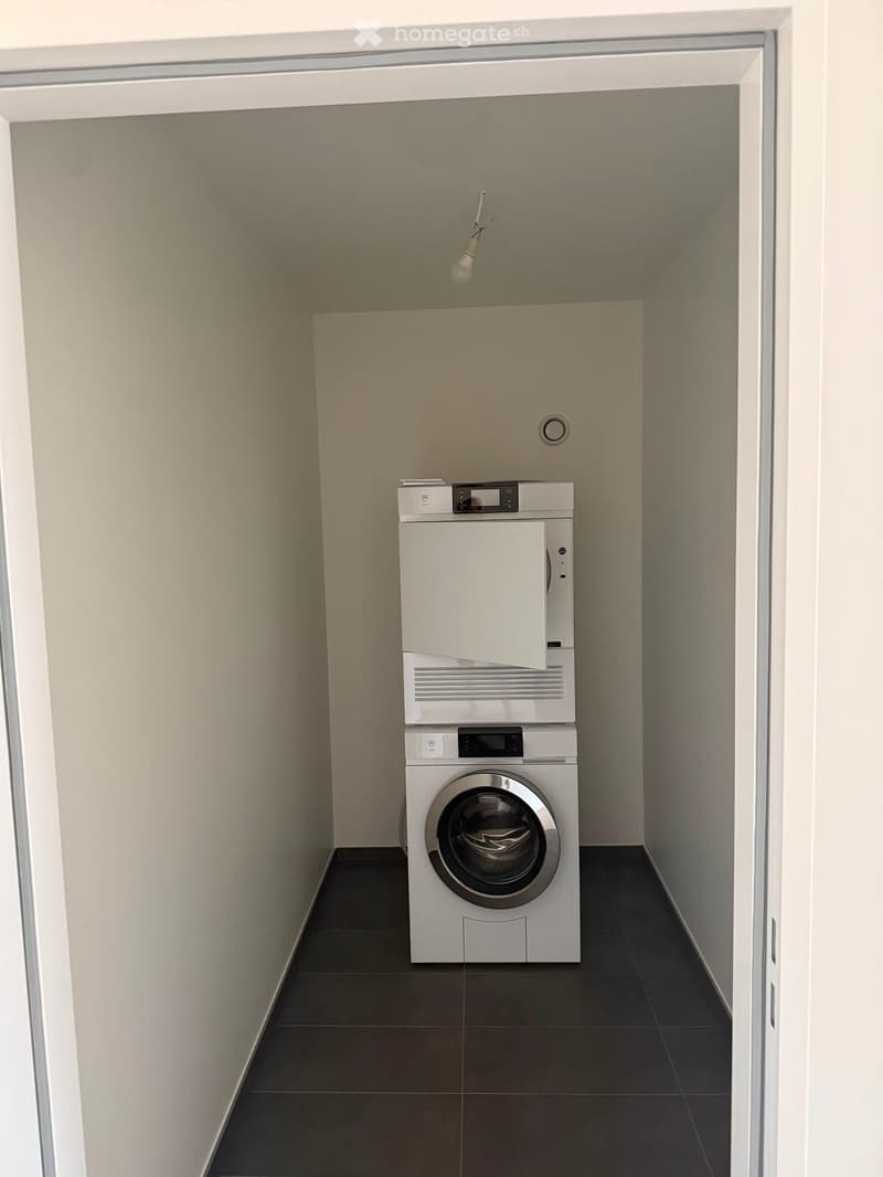 A narrow hallway with a washing machine and dryer stacked vertically, beige walls, and dark tiled flooring.