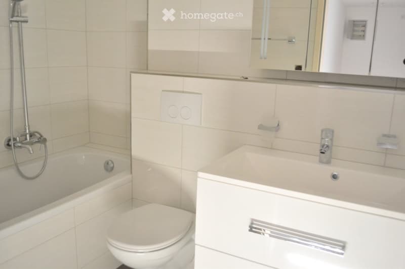 Modern bathroom with a bathtub, toilet, sink, mirror, and showerhead. The walls are tiled in light beige, and the fixtures are sleek and contemporary.