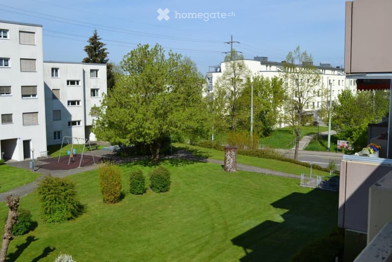A well-maintained garden area with green grass, small shrubs, and trees, surrounded by residential buildings and a playground.