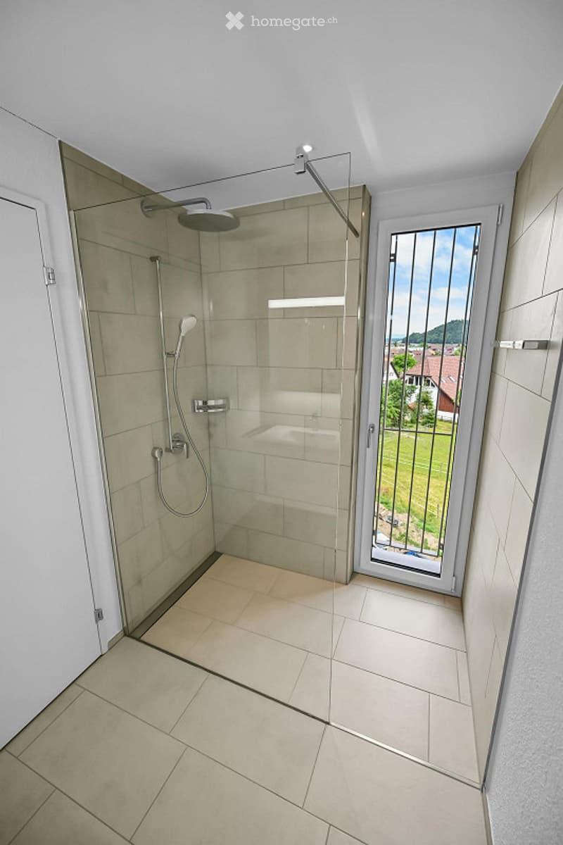 Modern bathroom with a walk-in shower, glass door, and tiled floor. Features a showerhead and faucet, with a view of the outdoor area through the glass door.