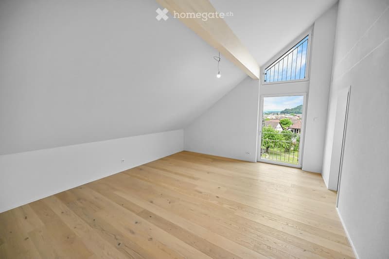Empty attic room with wooden flooring, white walls, and large windows offering a scenic view of the landscape.