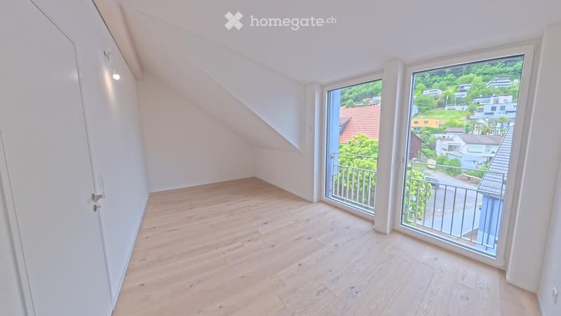 Bright, empty room with wooden flooring, white walls, and large windows offering a scenic view of the surrounding area.