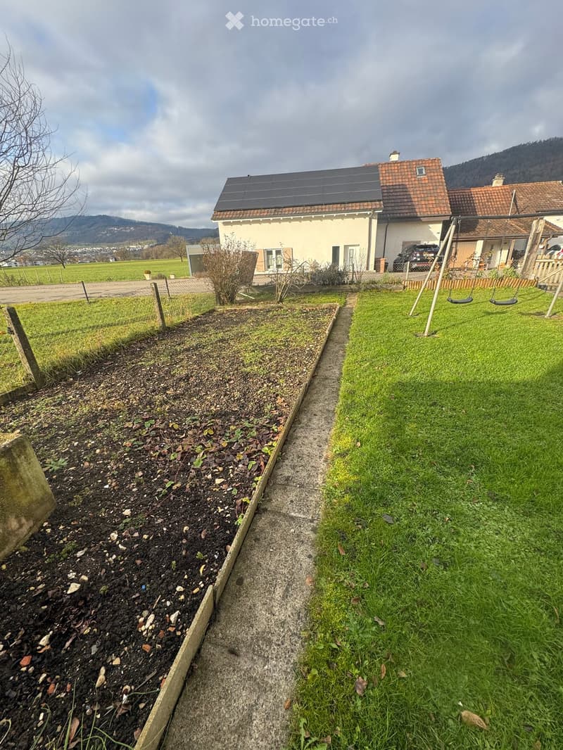 Garden area with a gravel path, raised garden bed, swing set, and a house with solar panels in the background.