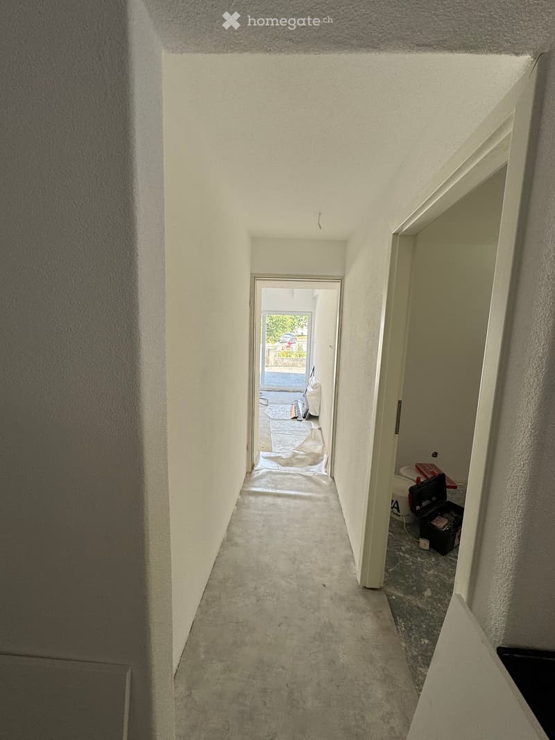 Empty hallway with white walls, concrete floor, and a doorway leading to an outdoor area. Some construction materials and tools are visible near the doorway.