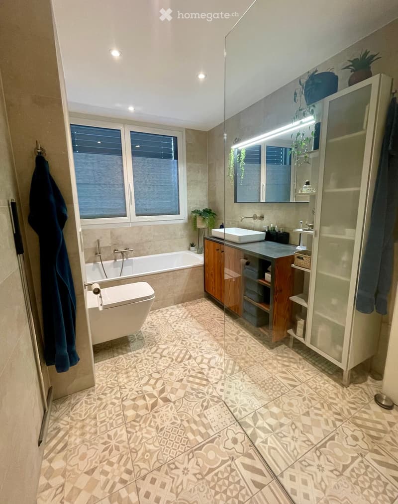 Modern bathroom with a bathtub, toilet, sink, and large mirror. Features wooden cabinetry, patterned tiles, and a glass-enclosed shower area.