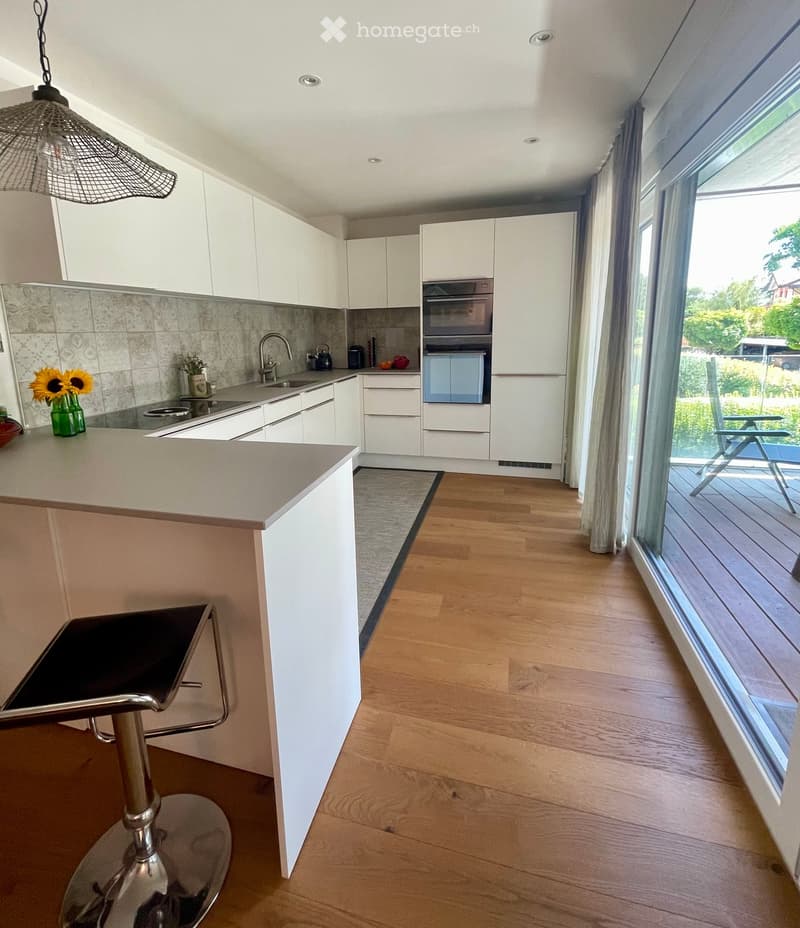Modern kitchen with white cabinets, wooden flooring, island with bar stool, sink, oven, and decorative vase on counter.