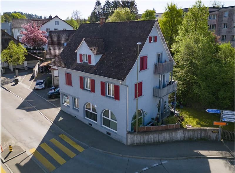 A two-story house with a dark roof and red shutters, surrounded by greenery and parked cars.