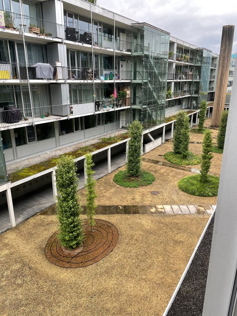 Courtyard area with gravel ground, small trees, and raised planters, surrounded by modern apartment building with balconies and glass railings.
